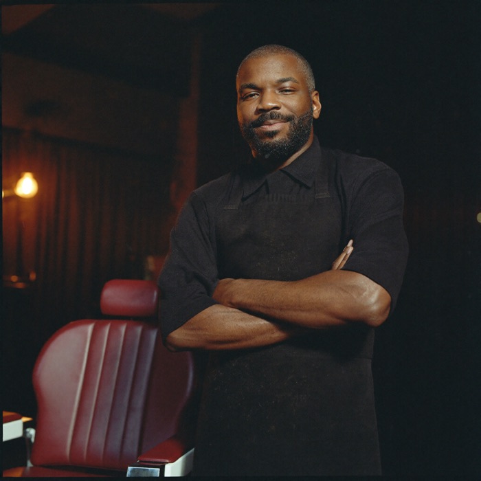 A master barber in a black apron standing beside a red leather chair.