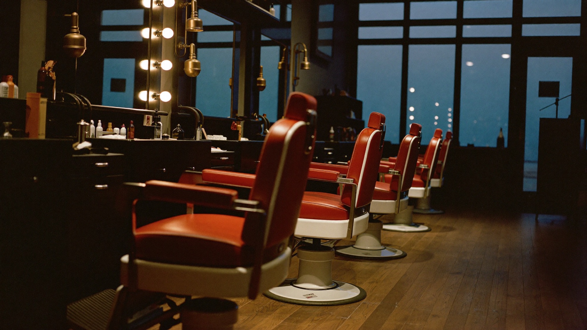 The C&C shop floor at dusk: a row of red leather barber chairs along black lacquer stations under warm bulbs.