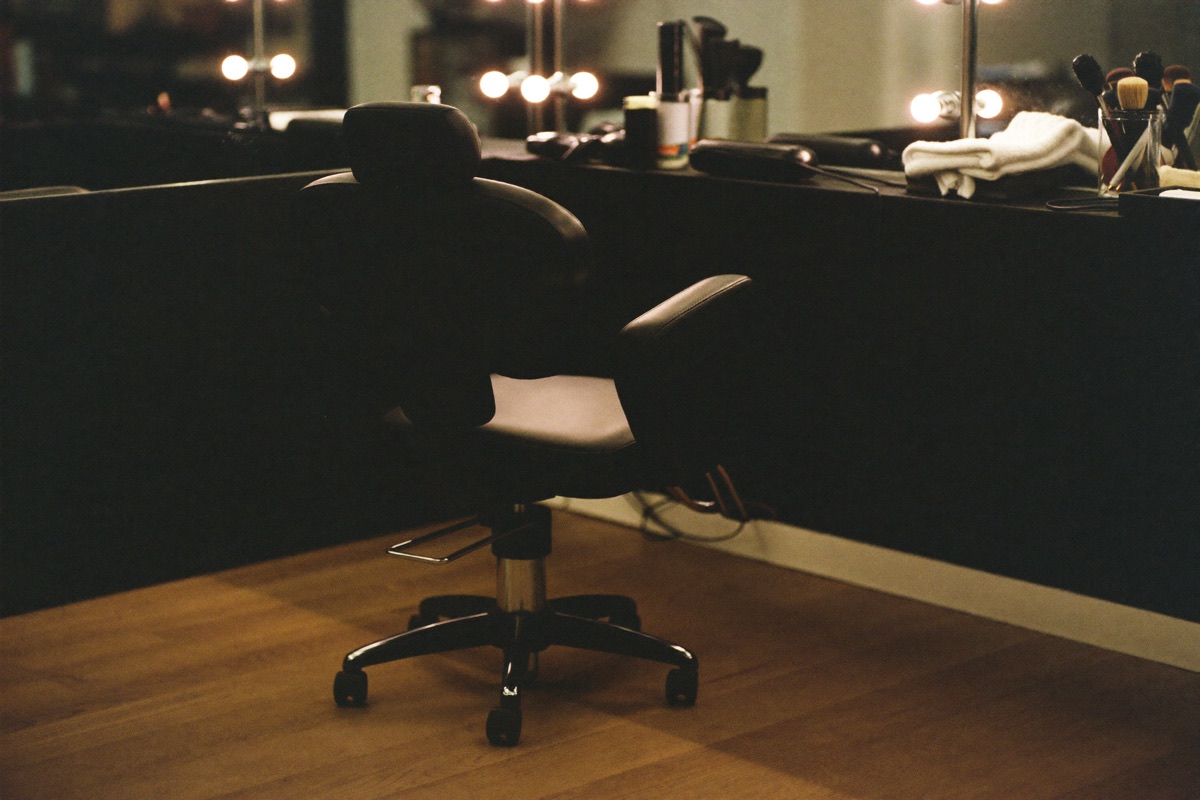 An empty salon styling chair in front of a bulb-lit mirror.